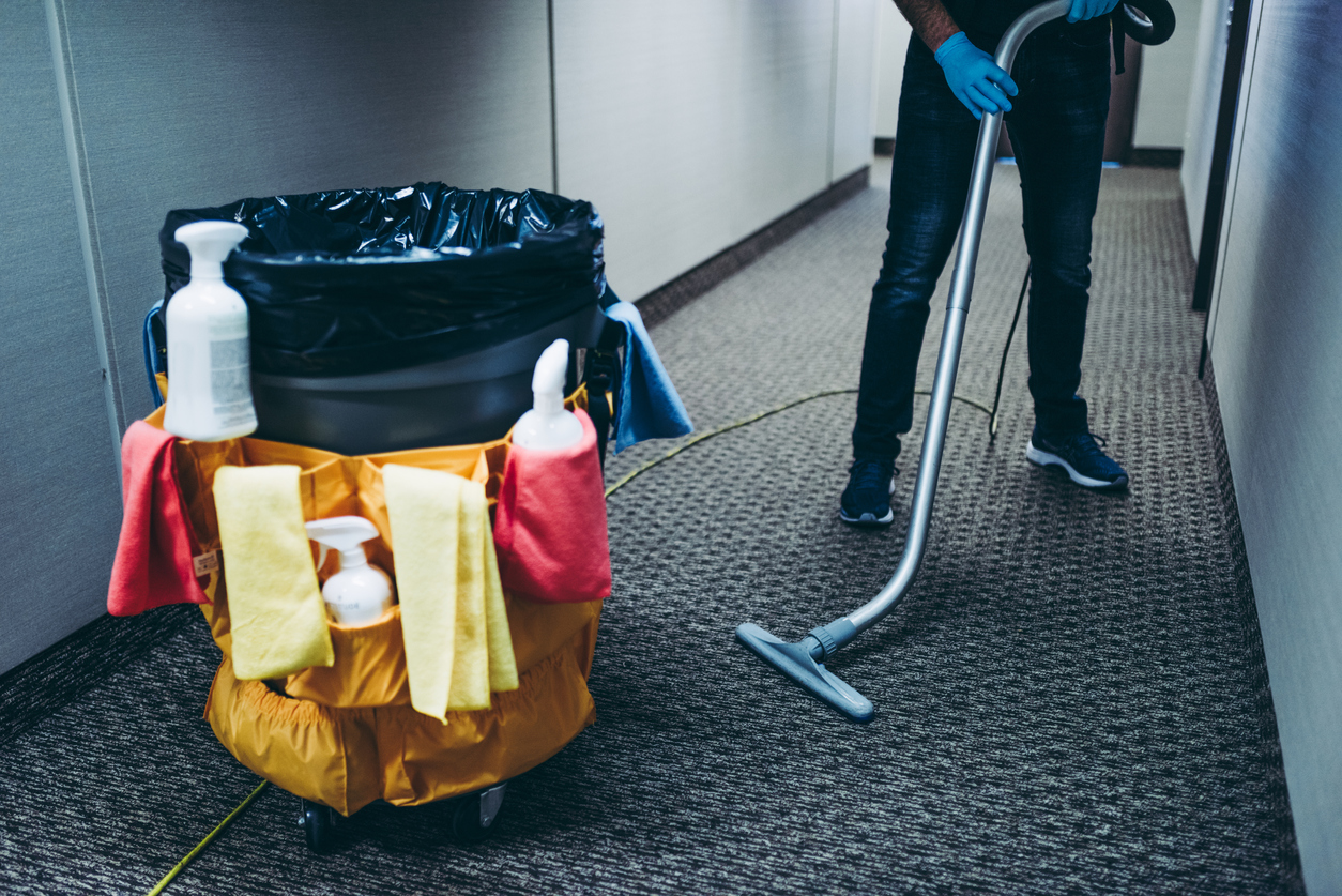 Janitor vacuum cleaning the corridor carpet floor inside an office building.around the garbage bin he has attached cleaning products.